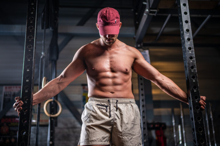 Shirtloze man met gespierd bovenlichaam, gekleed in beige sportshorts en een achterstevoren gedragen rode pet, leunend op een halterstang in een donkere gym setting. Hij kijkt omlaag met samengevouwen handen, in een moment van focus en kracht.